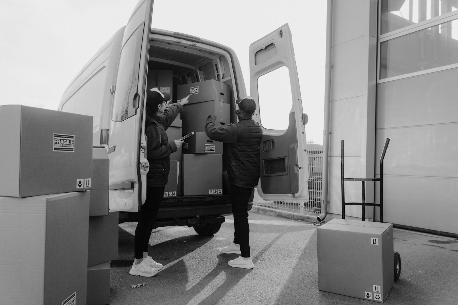 Two couriers loading cardboard boxes into a delivery van outdoors.